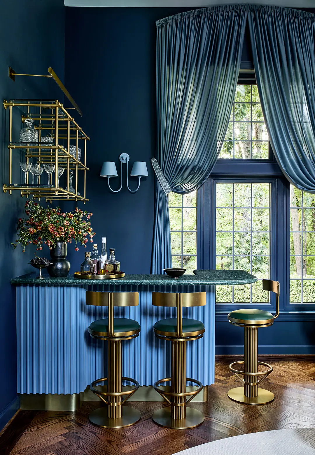 Bold blue bar room with deep navy walls, fluted blue cabinetry, brass detailing, and green leather stools, designed by Andrew Suvalsky Designs in Great Falls, Virginia