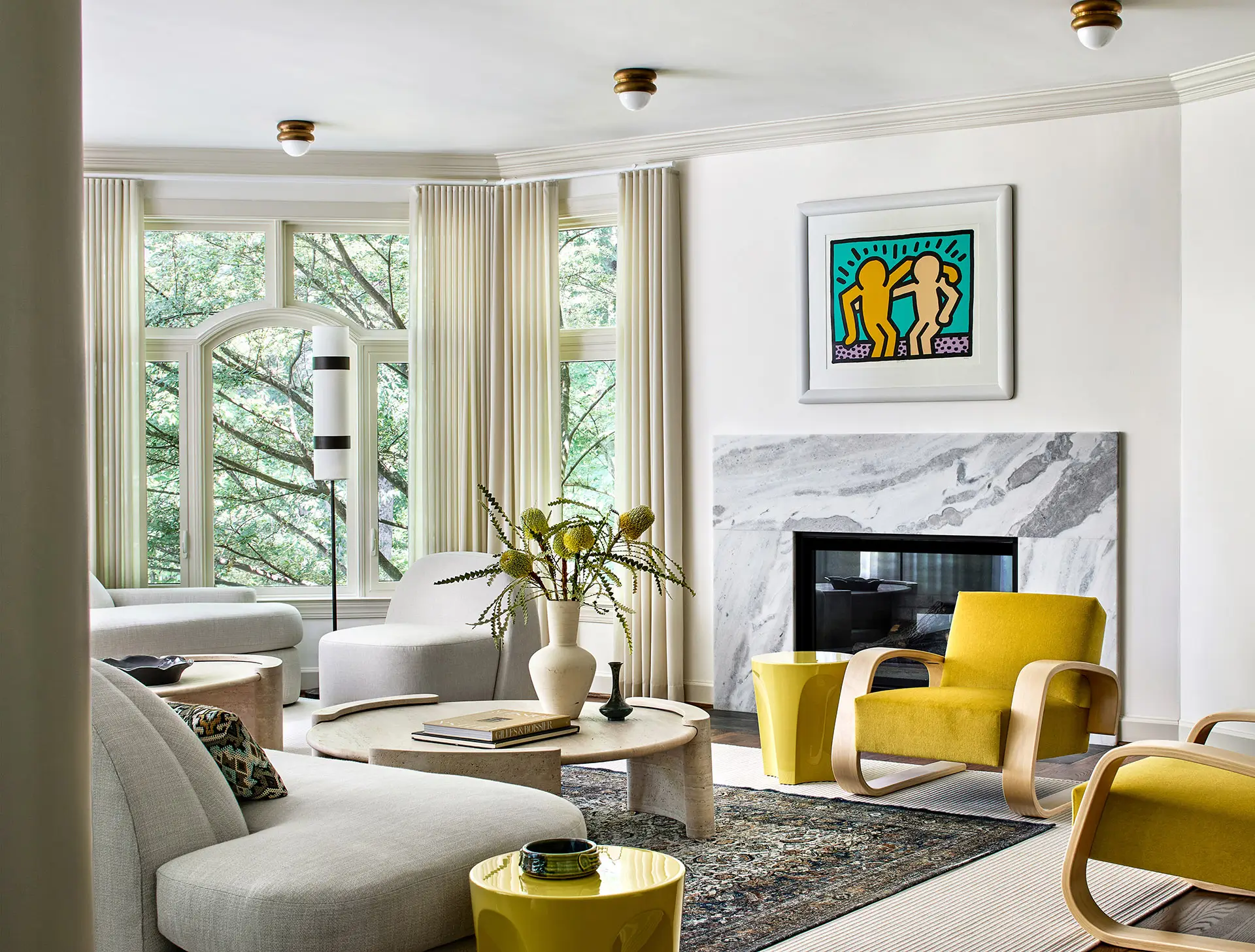 Great Falls, Virginia living room featuring framed Keith Haring artwork above a marble fireplace, with mustard yellow Alvar Aalto chairs and curved cream sofas, designed by Andrew Suvalsky Designs